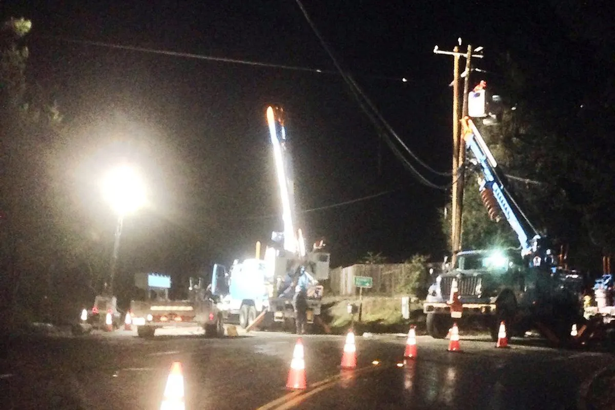 Line crews with bucket trucks working on power lines on a dark road at night, behind traffic cones